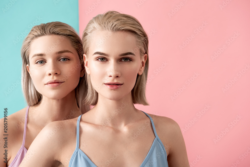 Portrait of young and well cared girls standing one after another back and staring at camera. Copy space in right side. Isolated on blue and pink background Portrait of young and well cared girls standing one after another back and staring at camera. Copy space in right side. Isolated on blue and pink background
