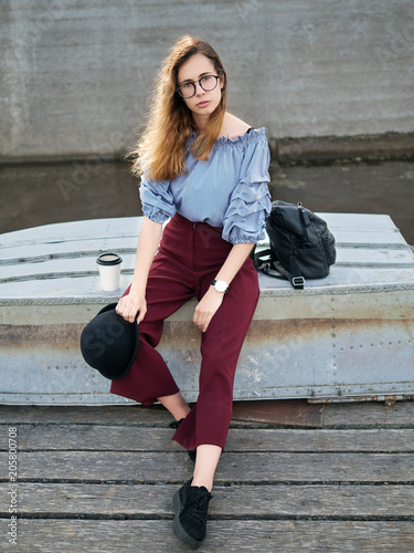 Young beautiful student girl in blue blouse and trendy glasses having fun with cup of coffee posing on a river pier in city park