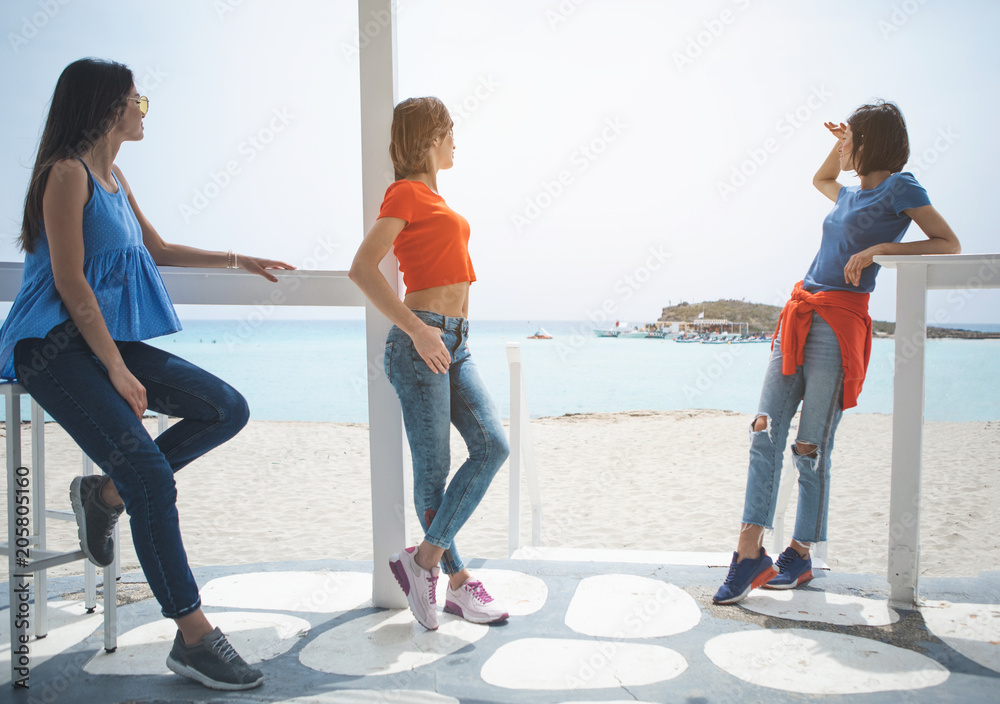 Fototapeta premium Carefree three girls are relaxing at the beach. They are standing inside the pavilion and looking at the sea with pleasure