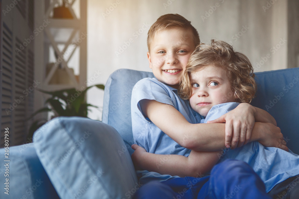 Portrait of happy brother embracing pensive kid while sitting on cozy seat in apartment Portrait of happy brother embracing pensive kid while sitting on cozy seat in apartment