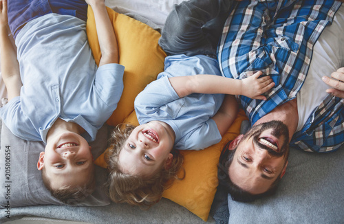 Portrait of cheerful bearded father relaxing on pillows with two little smiling kids. They looking at camera. Positive leisure at home concept
