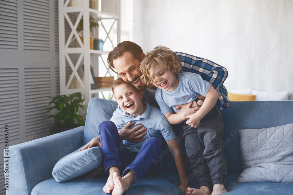 Portrait of laughing dad hugging beaming kids having entertainment on sofa in living room. Satisfied parent with glad children concept - obrazy, fototapety, plakaty 