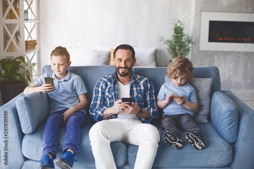 Portrait of cheerful bearded dad sitting between two smiling children on cozy sofa. They using gadgets. Happy family concept