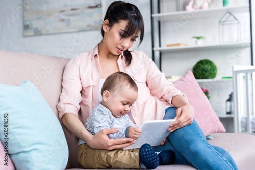 young happy mother and little child with tablet sitting on couch at home
