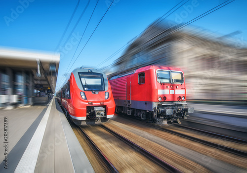 High speed train and old train in motion on the railway station at sunset. Modern intercity train on the railway platform. Industrial landscape. Commuter red trains with motion blur effect on railroad