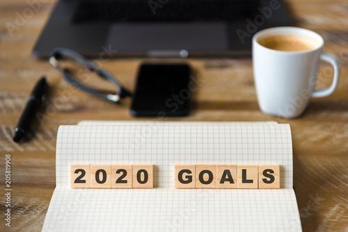 Closeup on notebook over wood table background, focus on wooden blocks with letters making 2020 Goals text