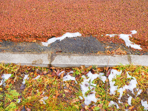 Top view Japanese park bike cycle lane smooth red asphalt with long cement block border and wet green, dry brown grass, black soil, fallen leaf, melting white snow pile dirt stains background