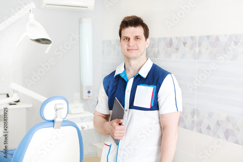 omatology and healthcare concept. Smiling male dentist  with tools directly into the camera over medical office background..