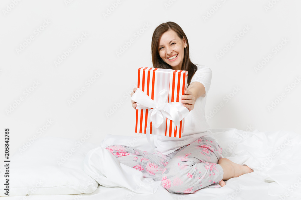 Young brunette woman sitting in bed with red striped gift box, white sheet, pillow, wrapping in blanket isolated on white background. Happy female spending time in room. Rest relax good mood concept. Young brunette woman sitting in bed with red striped gift box, white sheet, pillow, wrapping in blanket isolated on white background. Happy female spending time in room. Rest relax good mood concept.