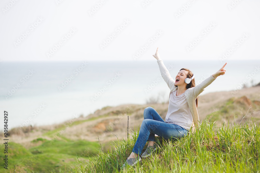 Young screaming fun pretty woman in casual clothes with headphones listening music sitting on grass spreading hands pointing index fingers up near water on green field background. Lifestyle concept. Young screaming fun pretty woman in casual clothes with headphones listening music sitting on grass spreading hands pointing index fingers up near water on green field background. Lifestyle concept.