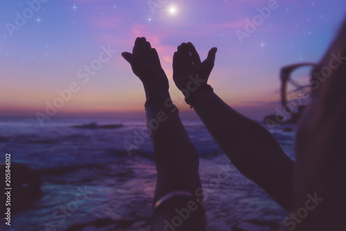 Silhouette of a girl with praying arms in dusk / dawn and glittering stars above the ocean. 