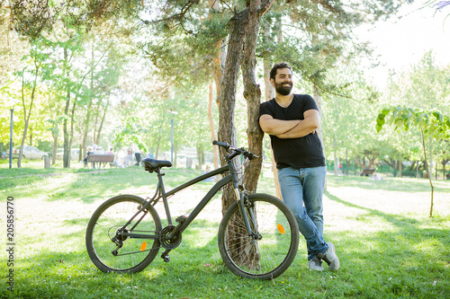 Smiling man leaning on a tree in the park with his bicycle next to him