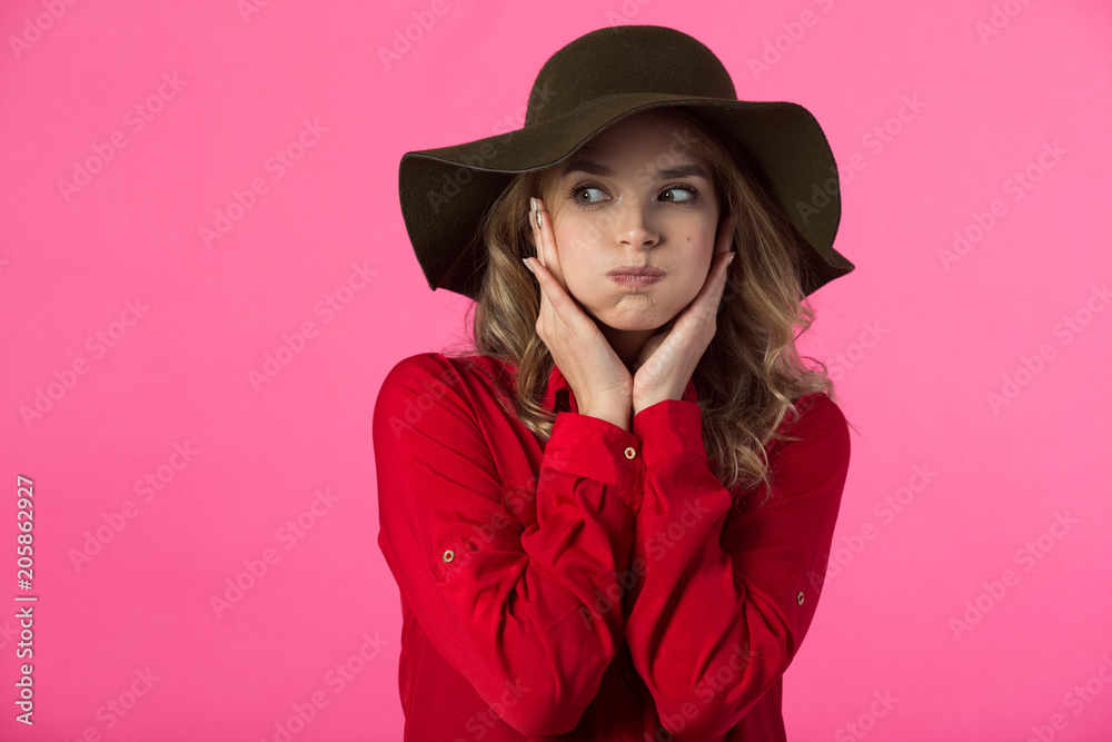 Fototapeta premium beautiful young girl in a red shirt and hat on a pink background with puffed cheeks