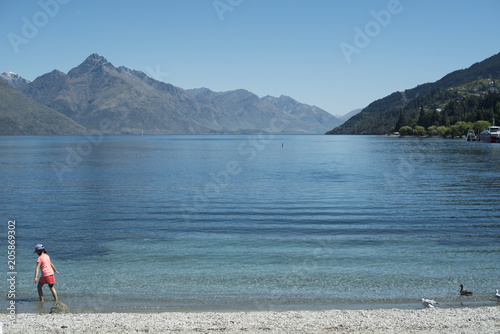 Personas descansando a la orilla de un lago de agua clara y azul frente a un paisaje de montañas Escena diurna, cielo azul y despejado. Nueva Zelanda.	