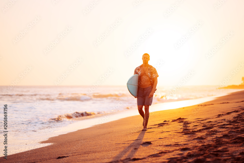 Male surfer walking on the beach near the ocean with short surfboard at sunset