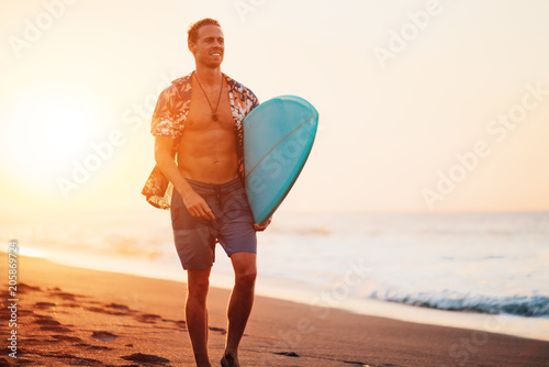 Guy surfer walking on the beach near the ocean with short surfboard at sunset