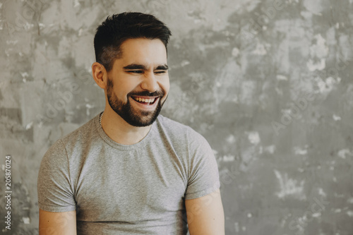 Portrait of a confident bearded man looking away and smiling closing one eye against a grey wall.