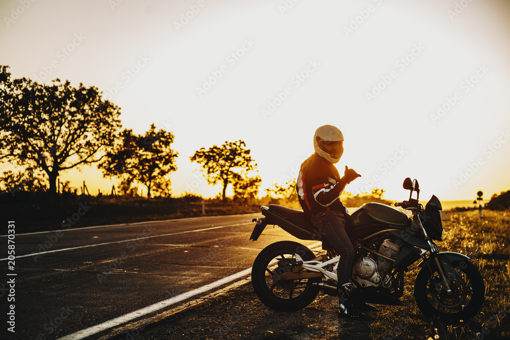 Fototapeta premium Amazing caucasian bike traveler sitting on his bike near the road and looking into camera showing that is ok against sunset.