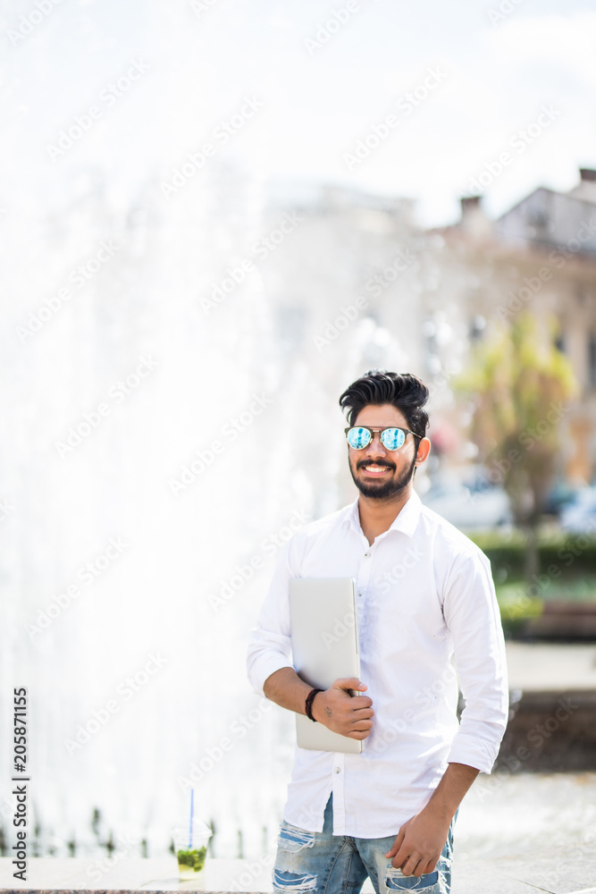 Portrait of a young serious indian man holding laptop computer while standing in a city area Portrait of a young serious indian man holding laptop computer while standing in a city area