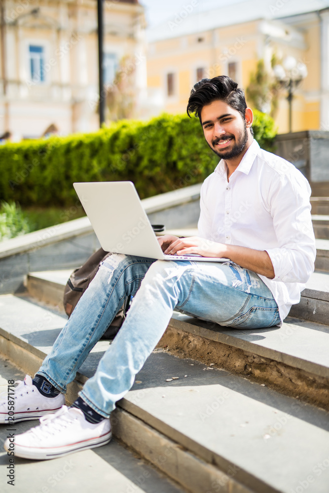 Handsome young indian manager working on laptop while sitting outdoors on the stairs in the street. Handsome young indian manager working on laptop while sitting outdoors on the stairs in the street.
