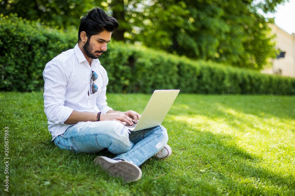 Young indian Student man with a laptop sitting on the grass. Urban style. Young indian Student man with a laptop sitting on the grass. Urban style.