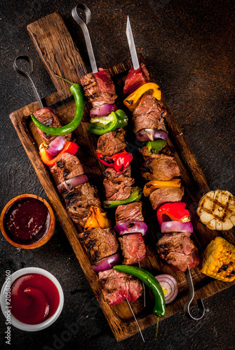 Fresh, home-cooked on the grill fire meat beef shish kebab with vegetables and spices, with barbecue sauce and ketchup, on a dark background on a wooden cutting board above copy space