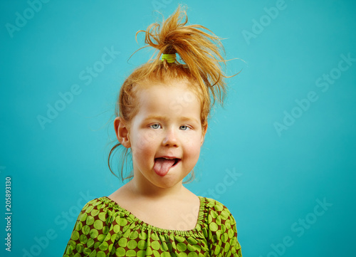 Εκτύπωση καμβά Close-up shot of beautiful squint girl three years old with tongue out, has red hair, is dressed in green dress, expresses a sweet sincerity, over blue isolated