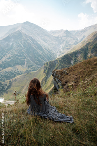 Beautiful young woman in a long dress is sitting on a cliff in the background of mountains