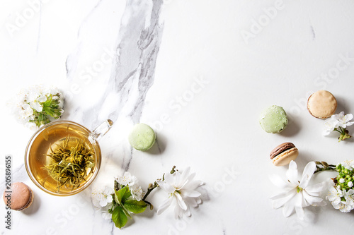 Glass cup of hot green tea with french dessert macaroons, spring flowers white magnolia and cherry blooming branches over white marble texture background. Top view, copy space.