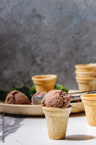 Homemade chocolate raspberry ice cream in small waffle cup served with frozen berries, fresh mint, chopped dark chocolate and metal spoon in ceramic plate on white marble kitchen table.