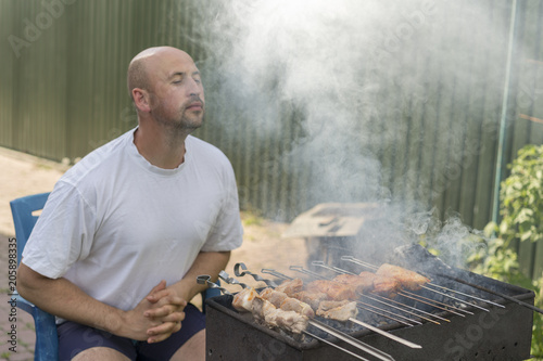 middle-aged man enjoys cooking meat on the grill. leisure, food, people and holidays concept - happy young man cooking meat on barbecue grill at outdoor summer party