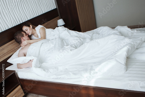 Young beautiful husband and wife resting in bed under a white blanket in a hotel room