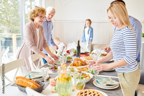 Portrait of happy two generation family serving festive dinner table together setting delicious homemade dishes for holiday  celebration in modern sunlit apartment
