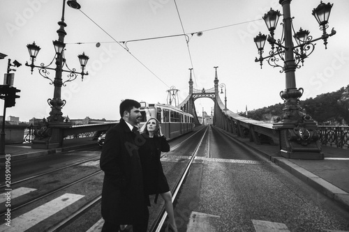 Couple in love is crossing the liberty bridge over the river Danube in Budapest. Man and woman in urban city. Vintage tram on bridge in Europe. Black and white stylish fashion photo in movement.