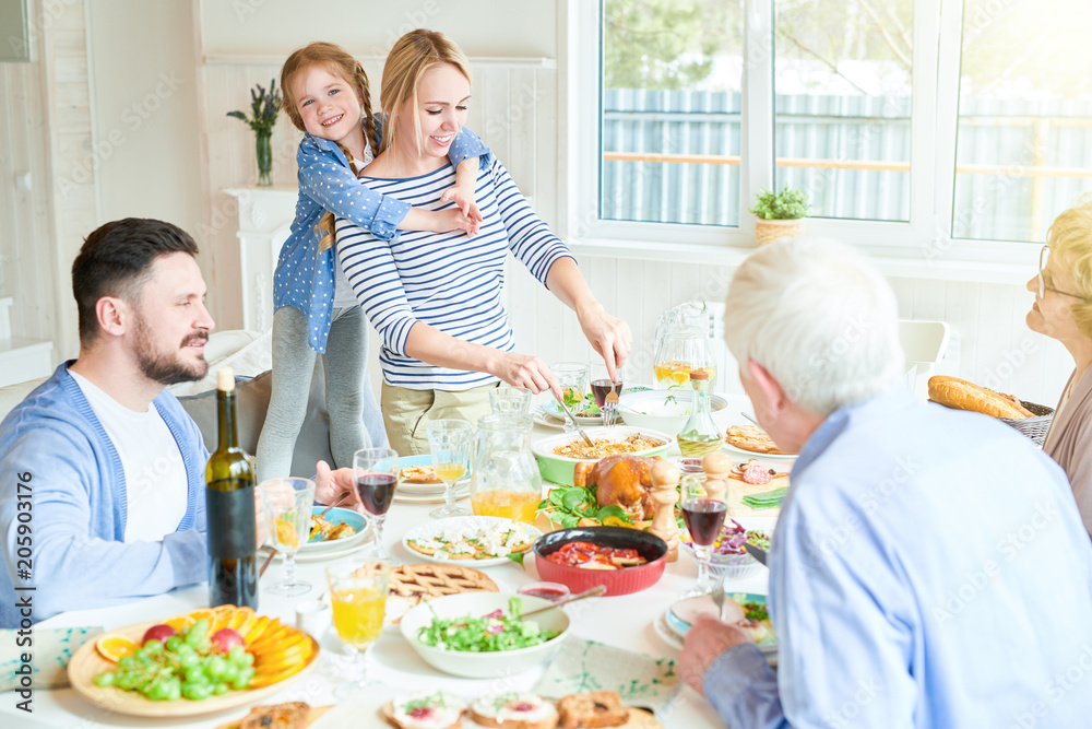 Portrait of happy family enjoying dinner sitting round festive table with delicious dishes, cute redhead girl hugging mom serving food during holiday celebration in modern sunlit apartment, copy space
