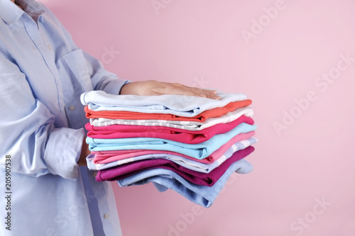 Close up of young woman wearing blue cotton shirt holding stack of perfectly folded multicolor shirts. Female w/ pile of different color clothing in her hands on pale pink background. Laundry concept.