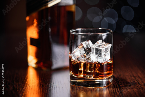 A glass of whiskey on the rocks and a bottle on the table on a dark background with a bokeh