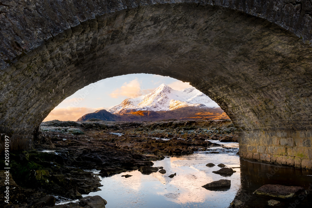 snow covered mountains framed by stone bridge arch at sunrise