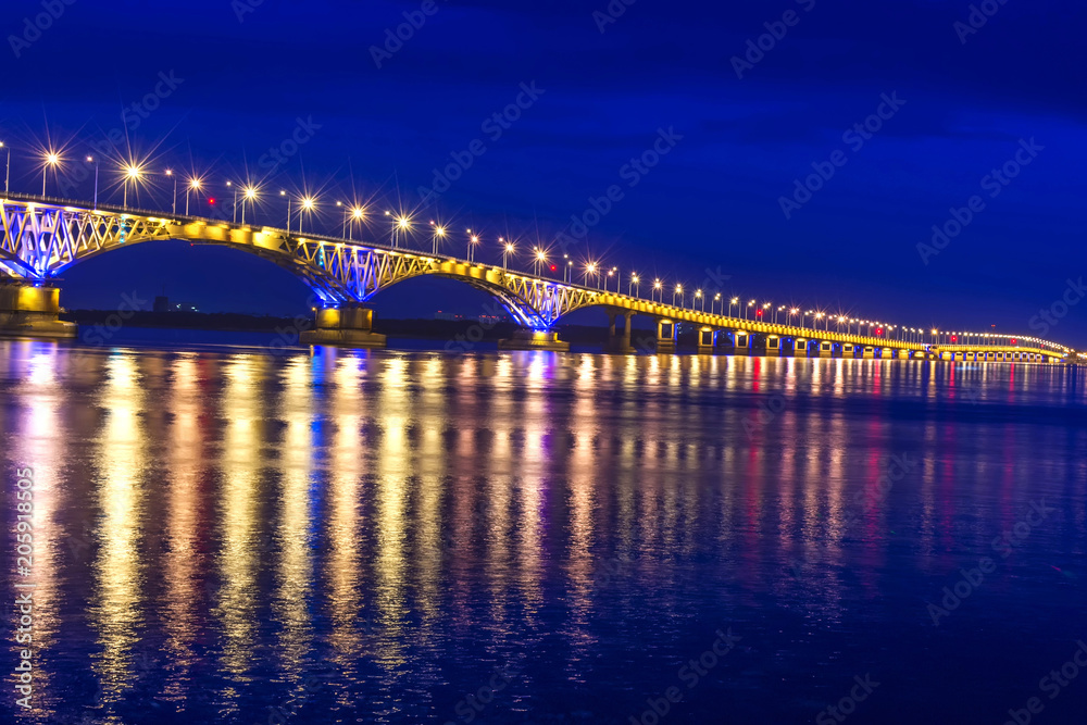 an automobile bridge across the Volga River at night, illuminated by the light of lanterns, the light of which is reflected in the river
