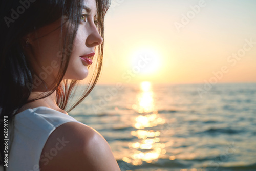 Close up of beatiful young girl looking on wonderful sea sunset. Having dark short hair. walking on sea waves. Looking on evening's deep blue sea . Amazing, colorful sunset. Wearing white shirt.
