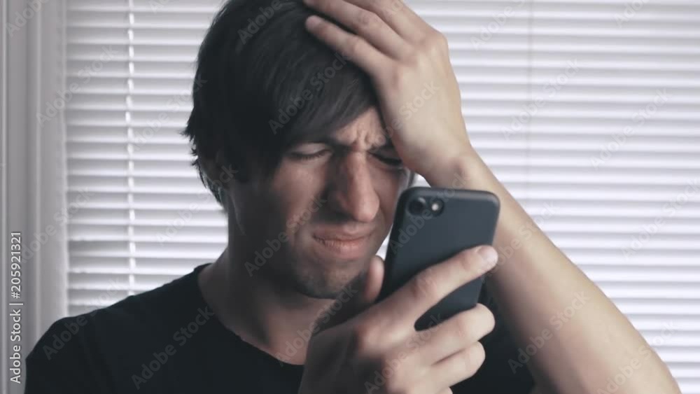 young man reading bad news on smartphone on the background of blinds in the office young man reading bad news on smartphone on the background of blinds in the office