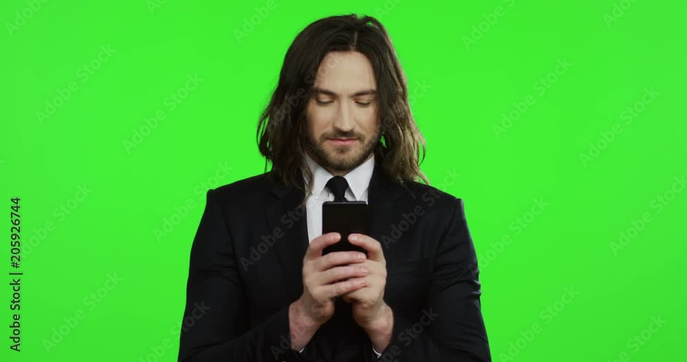 Portrait of the handsome young businessman in the black suit and tie scrolling and taping on the smartphone and then smiling on the chroma key background. Green screen. Portrait of the handsome young businessman in the black suit and tie scrolling and taping on the smartphone and then smiling on the chroma key background. Green screen.