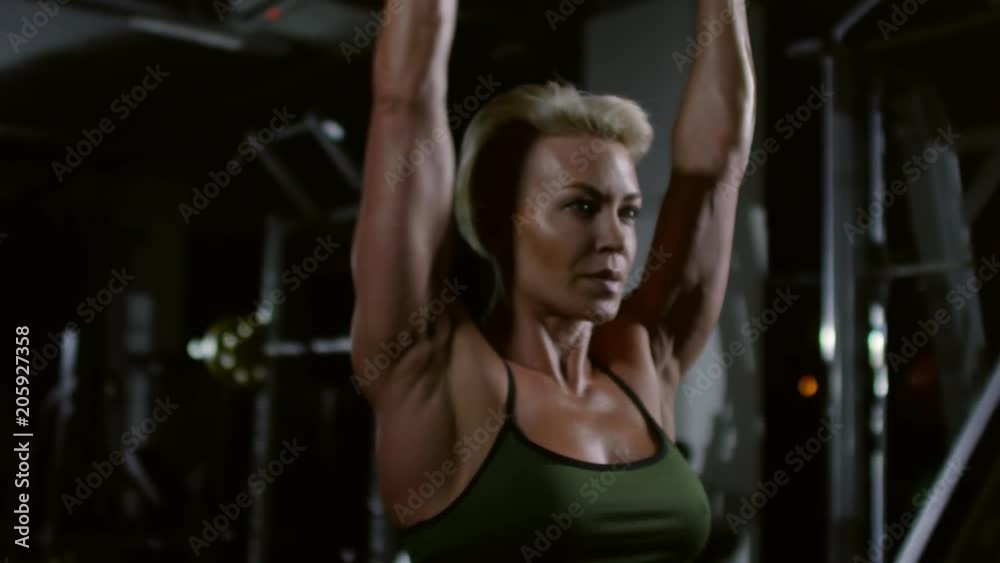 Attractive blonde woman with muscular arms doing dumbbell shoulder press while exercising in dark gym Attractive blonde woman with muscular arms doing dumbbell shoulder press while exercising in dark gym