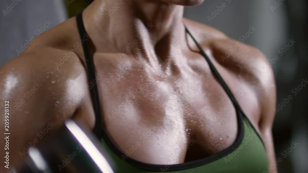 Closeup view of chest and shoulders of muscular woman with wet skin doing dumbbell curls