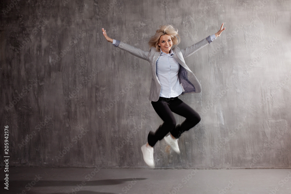Young blonde woman in business suit and sneakers jumping for joy, gray textured background