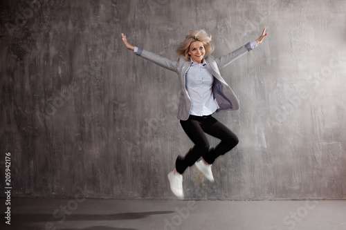 Young blonde woman in business suit and sneakers jumping for joy, gray textured background