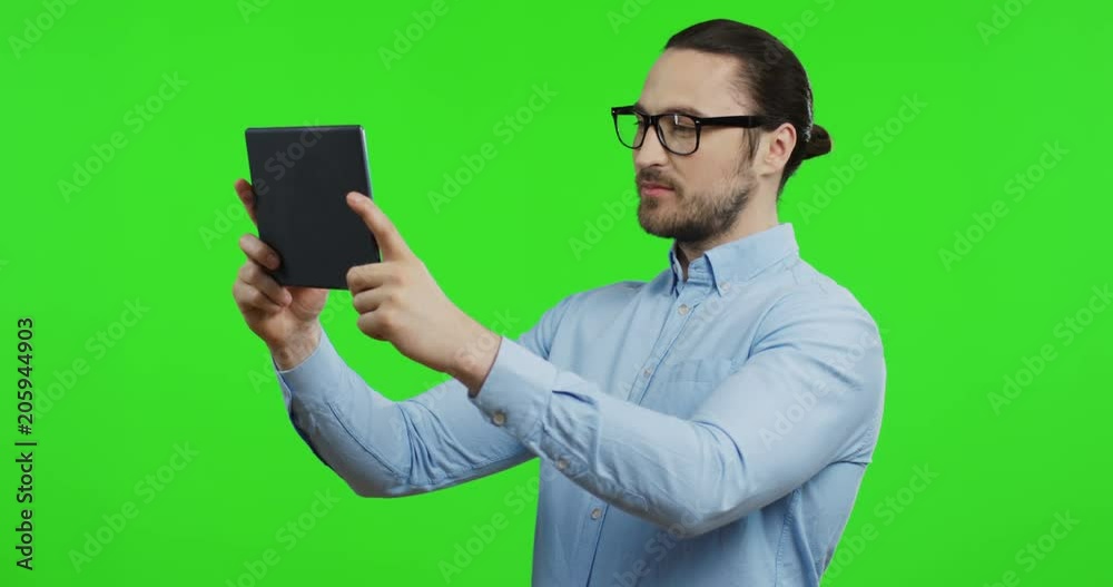 Good looking young man in glasses and blue shirt holding a tablet computer and watching something on it or doing selfie on the green screen background. Chroma key Good looking young man in glasses and blue shirt holding a tablet computer and watching something on it or doing selfie on the green screen background. Chroma key