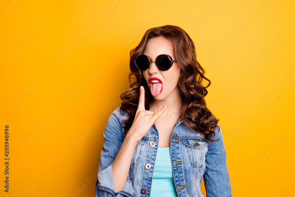 Portrait of crazy cool bitch fashionable girl in summer glasses gesturing tongue out showing rock and roll sign having modern hairdo isolated on yellow background