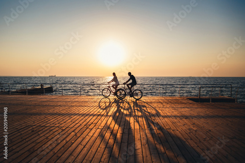 Couple of young hipsters cycling together at the beach at sunrise sky at wooden deck summer time