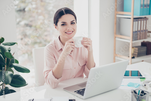 Portrait of cheerful charming trendy brunette classic woman enjoying break time sitting in modern white office with contemporary interior having mug with tea in hands looking at camera